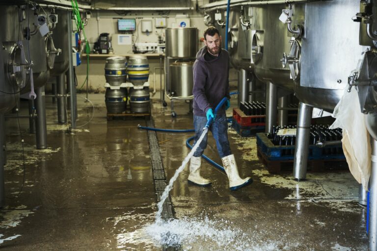 Man working in a brewery, cleaning floor with water hose.